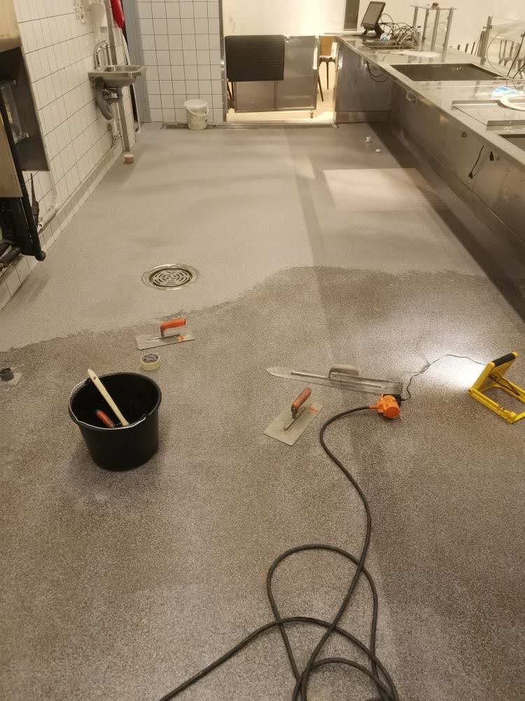 Keittiötilan lattialla on käynnissä lattiapinnoitustyö, jossa näkyy työkaluja ja osittain valmis pinnoite.